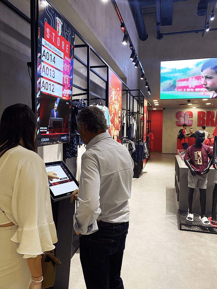 Two fans using the kiosk to get SMS tickets, choosing how to wait while shopping at SC Braga store.